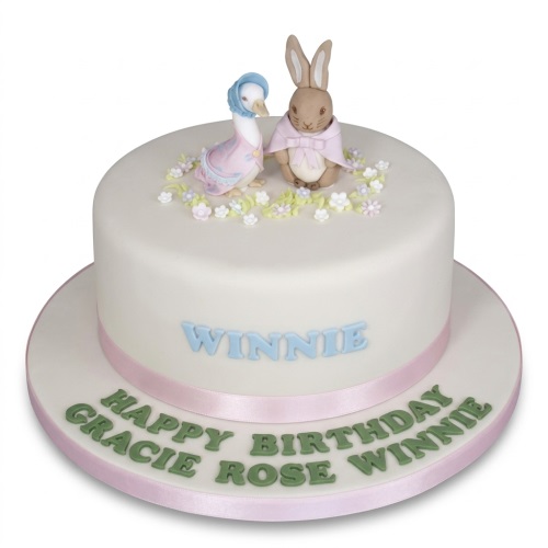 White round birthday cake with Peter Rabbit and Jemima Puddle-Duck figures, pastel flowers, and a pink ribbon.