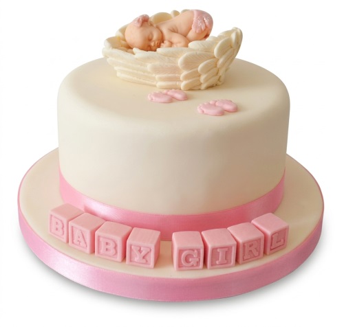 Round cream cake with pink ribbon, sleeping baby figurine in wings, and pink blocks spelling 'BABY GIRL'.