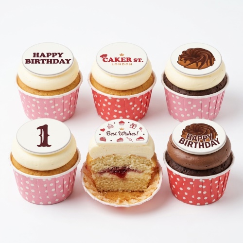 A selection of birthday cupcakes with white and brown frosting, pink and red polka dot wrappers, and custom edible toppers including 'Happy Birthday', 'Best Wishes!', and the number '1'.