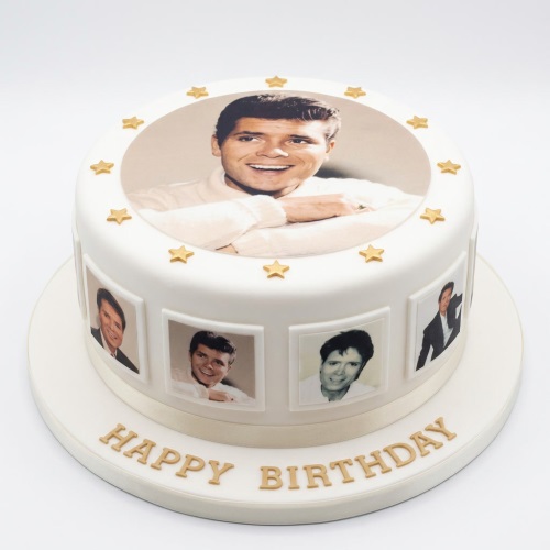 Round white birthday cake with a large edible photo on top and smaller photos around the side, decorated with gold stars and 'Happy Birthday' message.