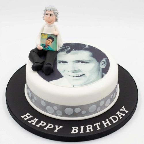 White round birthday cake with edible photo, sugar paste figurine holding a record, and grey polka dot ribbon.