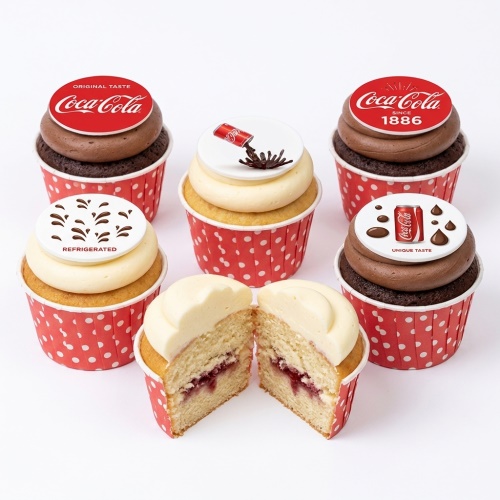 Six Coca-Cola themed cupcakes with red and white polka dot wrappers, featuring various Coca-Cola logo toppers and frosting designs. One cupcake is cut in half, showing a vanilla sponge with jam filling.