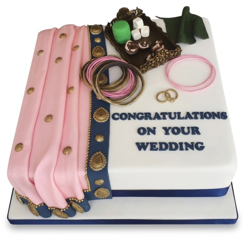 White wedding cake with pink saree drape, gold bangles, wedding rings, and sweets tray.