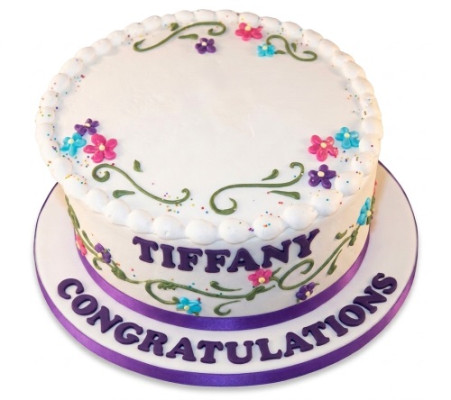 Round white celebration cake with colourful icing flowers, green vines, and a purple personalised message.