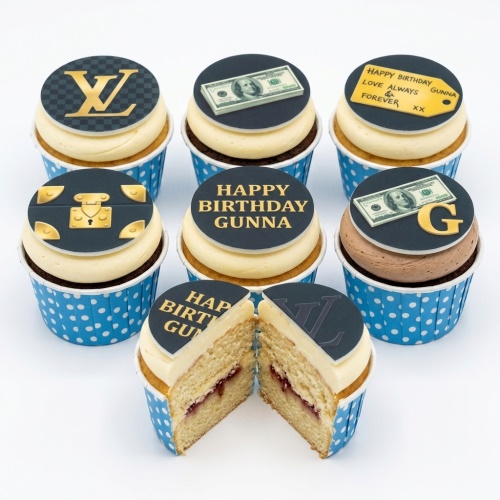 Cupcakes with luxury brand logo, money, and luggage trunk toppers on cream and brown frosting in blue polka dot cases.