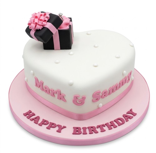 White heart-shaped birthday cake with pink ribbon, gift box with a ring, and personalised names.