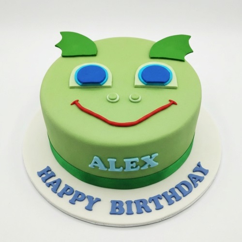 Green round birthday cake with a friendly dragon face, personalised with a name and 'Happy Birthday'.