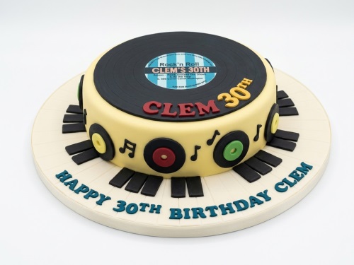 Round cream birthday cake with a black vinyl record on top and piano keys on the base, decorated with musical notes and small records.