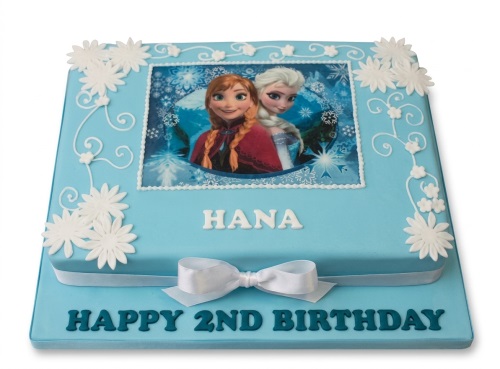 Light blue two-tiered square cake with an edible image of Anna and Elsa from Frozen, decorated with white sugar flowers and a white ribbon. The cake board has blue text 'Happy Birthday'.