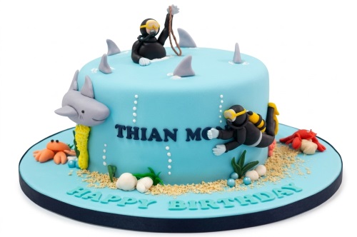 Round blue fondant cake with edible divers, shark fins, a shark, crabs, seashells, and sand decorations.