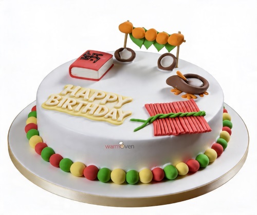 White round birthday cake with yellow "Happy Birthday" text, red firecrackers, orange toran, and diyas.
