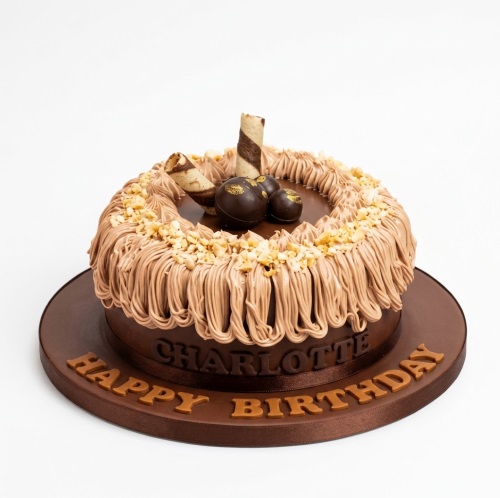 Round chocolate birthday cake with piped frosting, chopped nuts, chocolate spheres, and wafer rolls.