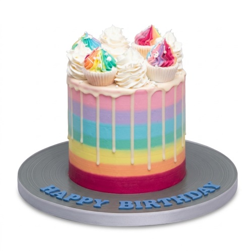Round rainbow drip birthday cake with white icing and mini rainbow cupcakes on top. Blue "Happy Birthday" message on the board.