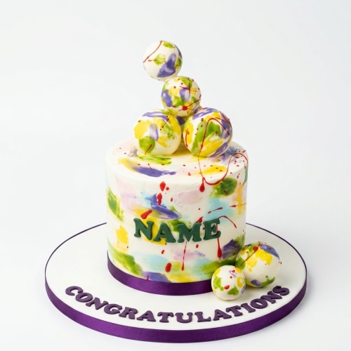 Round white cake with colourful paint splatters, decorative spheres, and a purple ribbon.