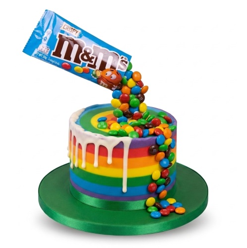 Round rainbow cake with white drip and M&M's pouring from a blue M&M's crispy bag.