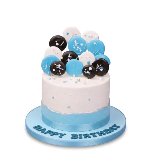 Round white and blue birthday cake with black, white, and blue confetti-style toppers and edible pearls.
