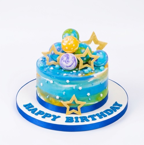 Round blue galaxy birthday cake with colourful planets, gold stars, and white sprinkles.