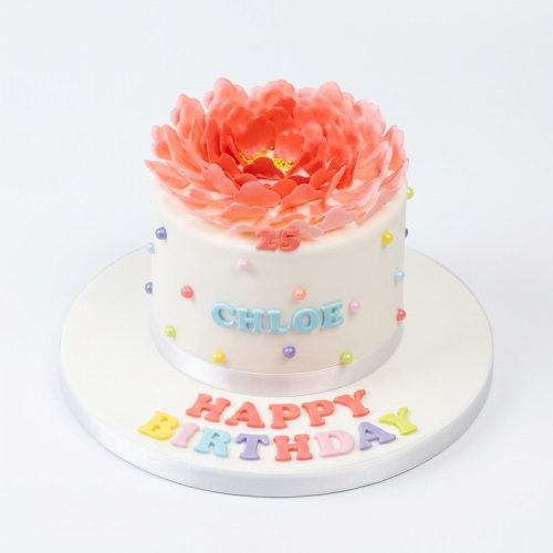White single-tier floral birthday cake with large coral flower, pastel sprinkles, and custom age.
