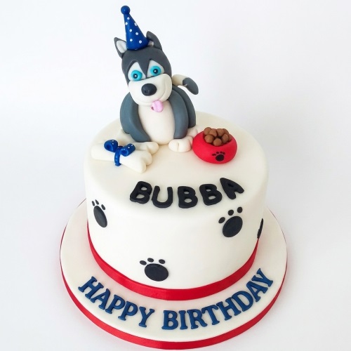White fondant birthday cake with a grey husky dog figure, blue party hat, red dog bowl, bone, and black paw prints.