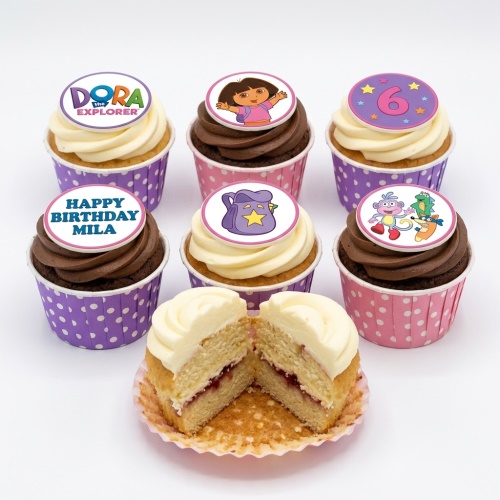 Dora the Explorer themed cupcakes with white and chocolate frosting, featuring edible toppers of Dora, her backpack, and friends, with a custom age '6' and 'Happy Birthday Mila' message.