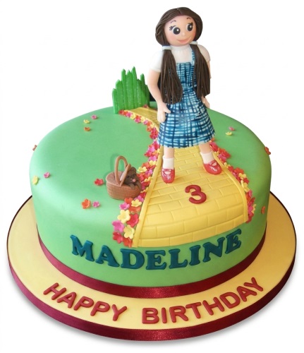Round green cake with Dorothy figure, yellow brick road, basket with dog, and colourful flowers.