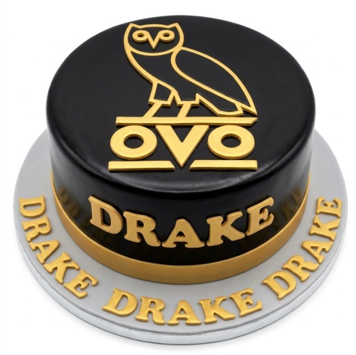 Round black cake with gold OVO owl logo and 'DRAKE' lettering, on a grey board.