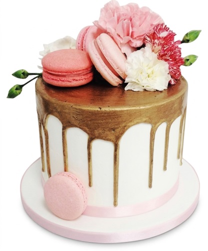White cake with gold drip, pink and white flowers, and pink macarons.