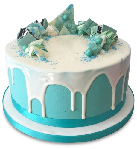 Round blue drip cake with white icing, blue and white chocolate shards, meringues, and sprinkles on a white cake board with a blue ribbon.