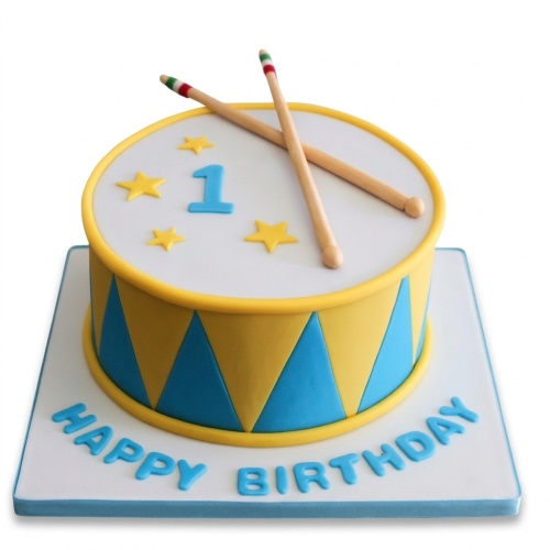 Round drum theme birthday cake with yellow and blue sides, white top, drumsticks, stars, and number one topper.
