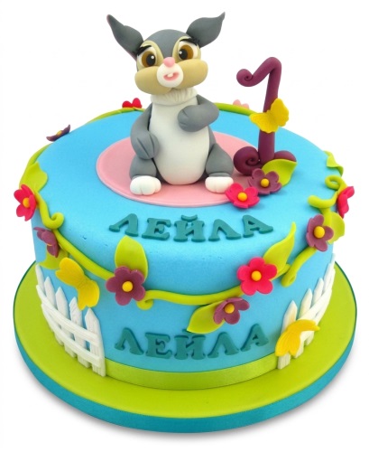 Round blue 1st birthday cake with a 3D grey and white bunny, number one topper, pink and purple fondant flowers, green vines, and a white picket fence.