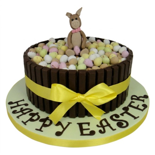 Round Easter cake with chocolate fingers, mini eggs, a bunny topper, and a yellow ribbon.