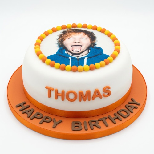 Round white photo birthday cake with edible image of Ed Sheeran, orange and yellow beads, and orange board.