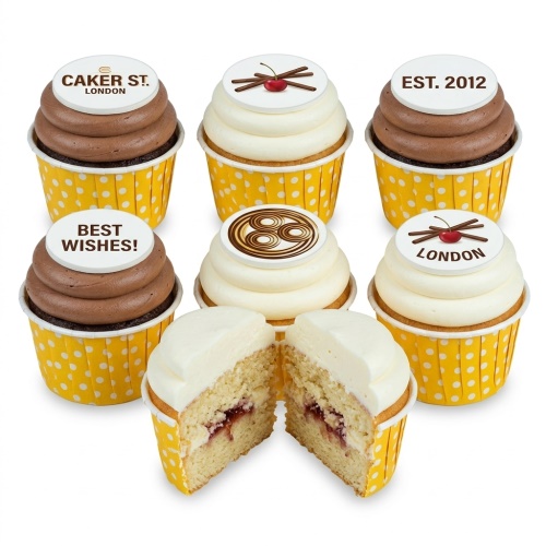 Cupcakes with yellow polka dot wrappers, white and chocolate frosting, and edible toppers featuring 'Best Wishes', 'Caker St. London', and a cherry design. One cupcake is cut open showing vanilla sponge with jam filling.