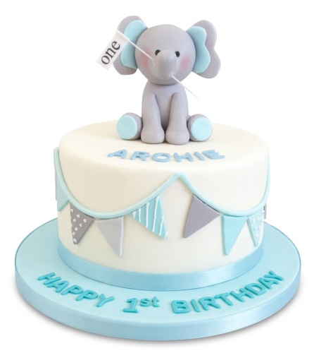 White 1st birthday cake with a grey elephant topper holding a 'one' flag, light blue and grey bunting, and a light blue ribbon.