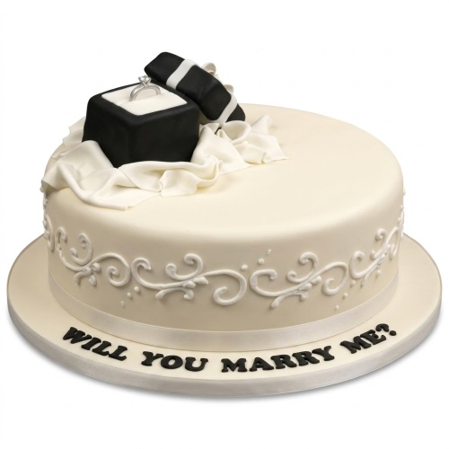 White round cake with a black ring box, silver ring, and 'Will You Marry Me?' message.