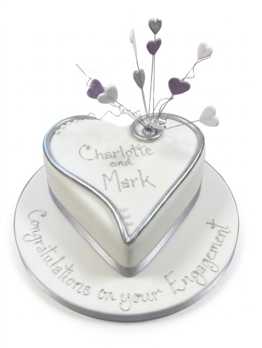 White heart-shaped engagement cake with silver and lilac hearts, silver rings, and customisable names.