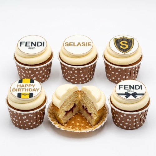 Six Fendi inspired cupcakes with white frosting, brown polka dot cases, and edible designer logo toppers for a birthday celebration.