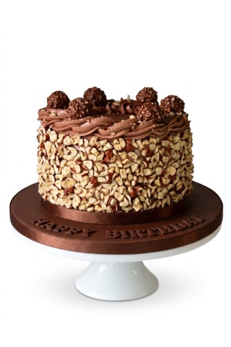 Round chocolate cake covered in chopped hazelnuts, topped with chocolate cream swirls and chocolate hazelnut spheres, on a brown cake board with "Happy Birthday" message.