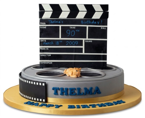 Two-tier movie theme birthday cake with clapperboard topper, film reel base, and small cat figurine.