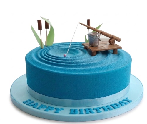 Round blue fishing theme birthday cake with edible dock, fishing rod, bucket, and cattails.