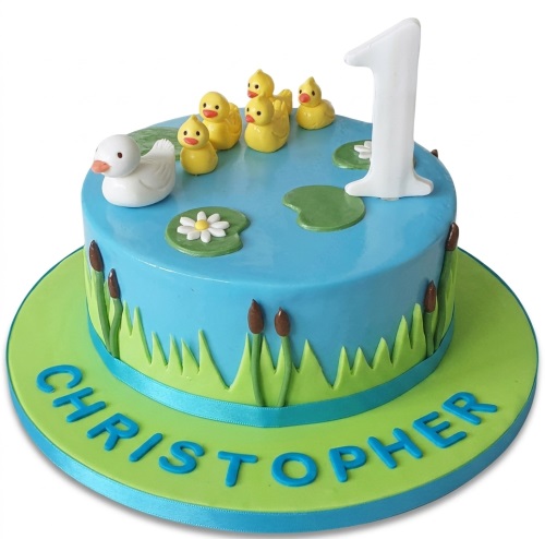 Round blue and green duck pond birthday cake with yellow ducklings, a white duck, lily pads, reeds, and a white number topper.