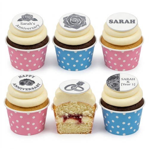 Six anniversary cupcakes with white frosting, rose and wedding ring toppers, in pink and blue polka dot cases.