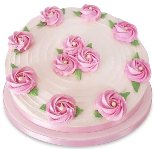 Round pink birthday cake with light pink buttercream, decorated with darker pink buttercream roses, green leaves, and pearl accents, on a pink cake board with a ribbon.