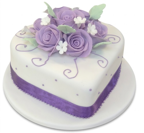 White heart shaped cake with purple sugar roses, green leaves, and a purple ribbon.