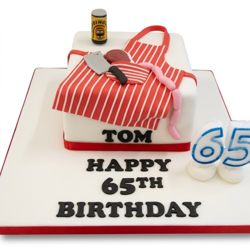 White square birthday cake with a red and white striped butcher's apron, cleaver, sausages, and a tin can.