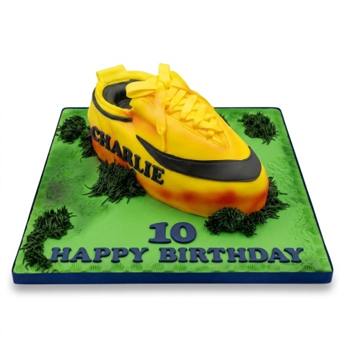 3D yellow and orange football boot birthday cake on a green pitch board with custom age and message.