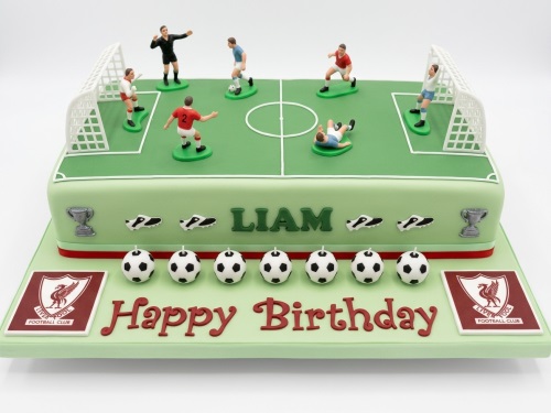 Rectangular green football pitch cake with player figurines, goals, footballs, and Liverpool FC crests.