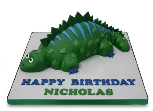 3D green dinosaur birthday cake with blue details and a personalised message on a white board.
