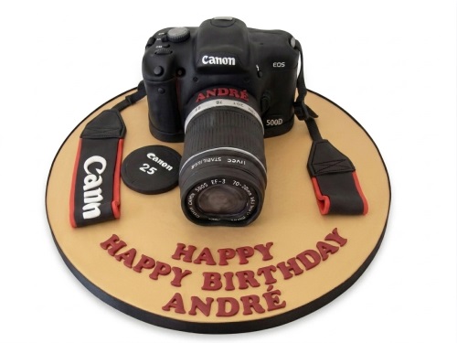 A 3D sculpted black camera cake with a red and black strap, on a tan cake board with a 'Happy Birthday' message.