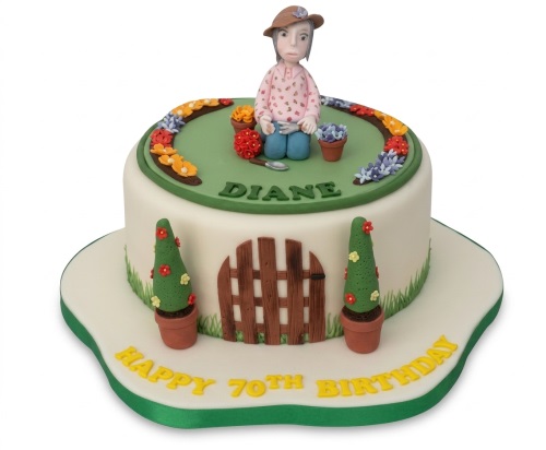 Round white birthday cake with a gardening theme, featuring a female gardener figure, flowers, fence, and topiary trees.